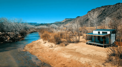 A small cabin by a river, surrounded by dry grass and bare trees, with rocky hills in the background under a clear blue sky.